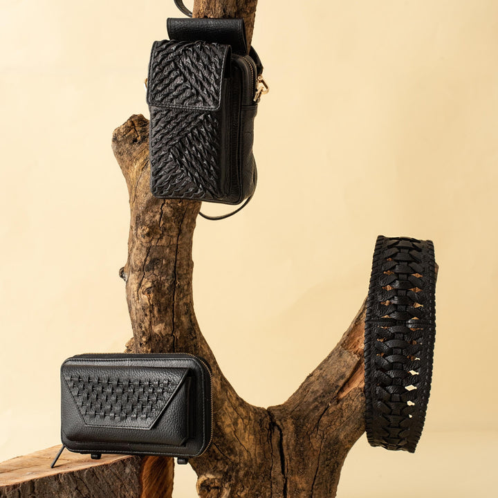 Black woven leather crossbody bag, wallet, and belt displayed on rustic tree branch with beige background