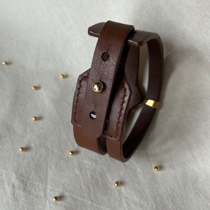 Brown leather bracelet with metal stud and gold accent on white fabric background with scattered small gold beads