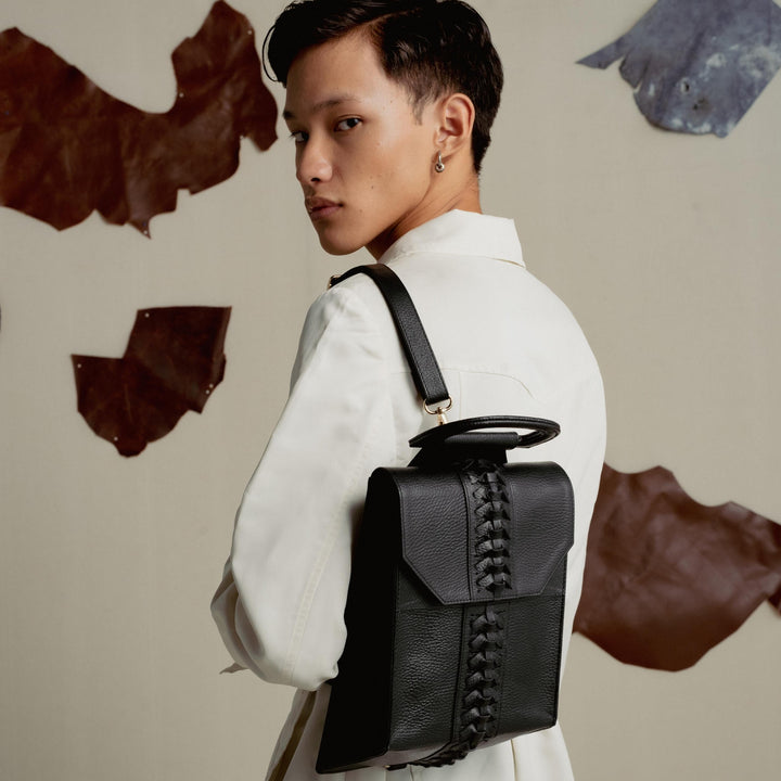 Young person in white jacket with black leather backpack featuring woven detail, neutral background with leather patches