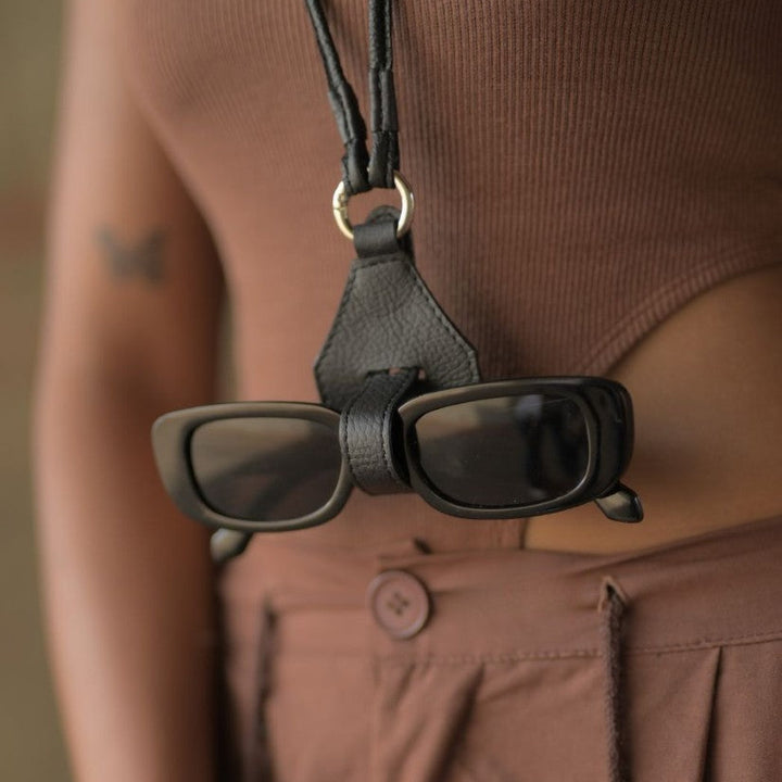 Black rectangular sunglasses secured on a black leather protective case hanging from neck on a person wearing brown top and pants